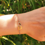 Gros plan d'un délicat bracelet chaine en métal bicolore, avec deux coeurs évidés entrelacés. L'un est de couleur argent, l'autre de couleur or jaune, reposant sur un poignet avec un arrière-plan flou de feuillage vert ensoleillé.