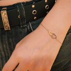 Vue à mi-corps d'une personne portant un bracelet chaine délicat avec deux coeurs entrelacés bicolores (argent et or) à son poignet. La main repose sur le devant d'un jean en jean gris foncé, à côté d'une ceinture en cuir noir avec une boucle gravée ornée.