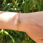 Un plan très rapproché et détaillé de la section centrale d'un bracelet chaine, montrant précisément l'entrelacement de deux breloques en forme de coeur évidées : un coeur argenté bicolore et un coeur doré bicolore, maintenus par des anneaux de saut sur une chaine en métal argenté. L'arrière-plan est une verdure ensoleillée et floue.