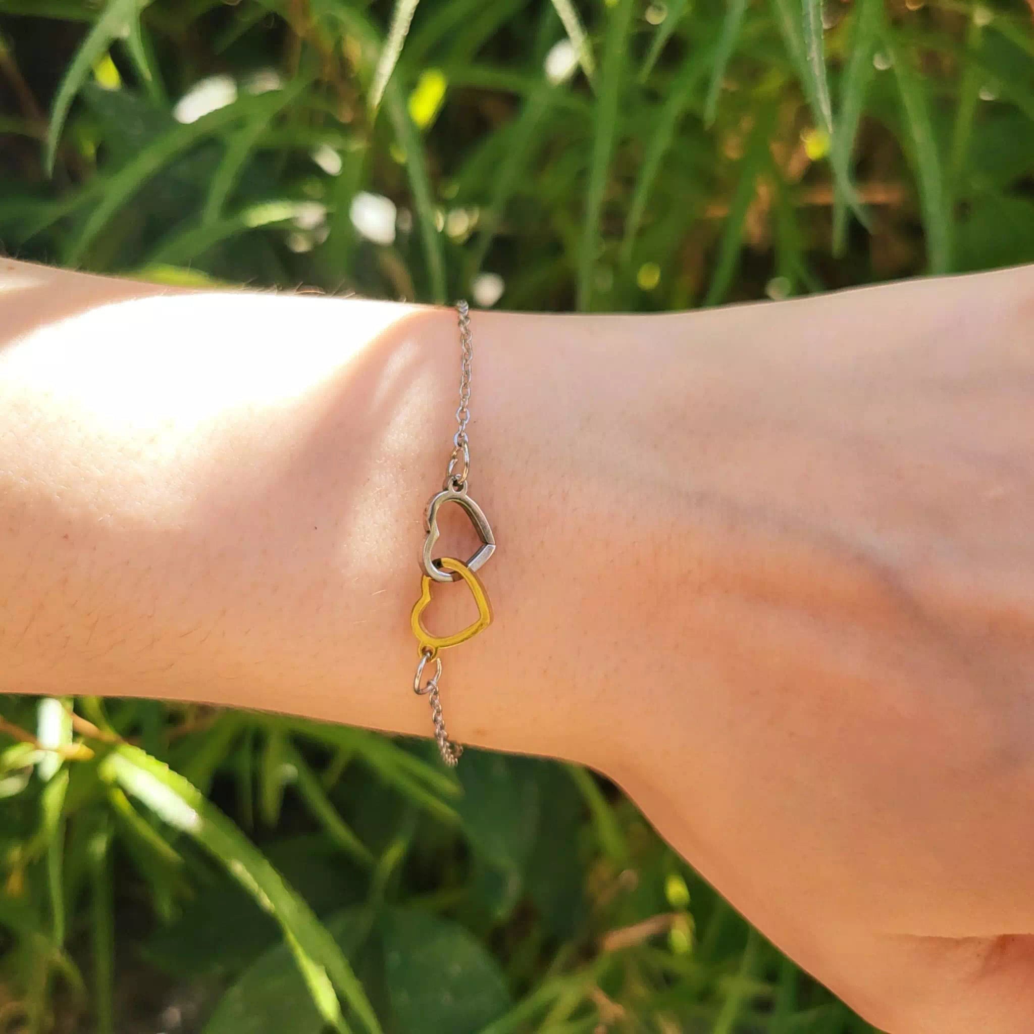 Un plan très rapproché et détaillé de la section centrale d'un bracelet chaine, montrant précisément l'entrelacement de deux breloques en forme de coeur évidées : un coeur argenté bicolore et un coeur doré bicolore, maintenus par des anneaux de saut sur une chaine en métal argenté. L'arrière-plan est une verdure ensoleillée et floue.