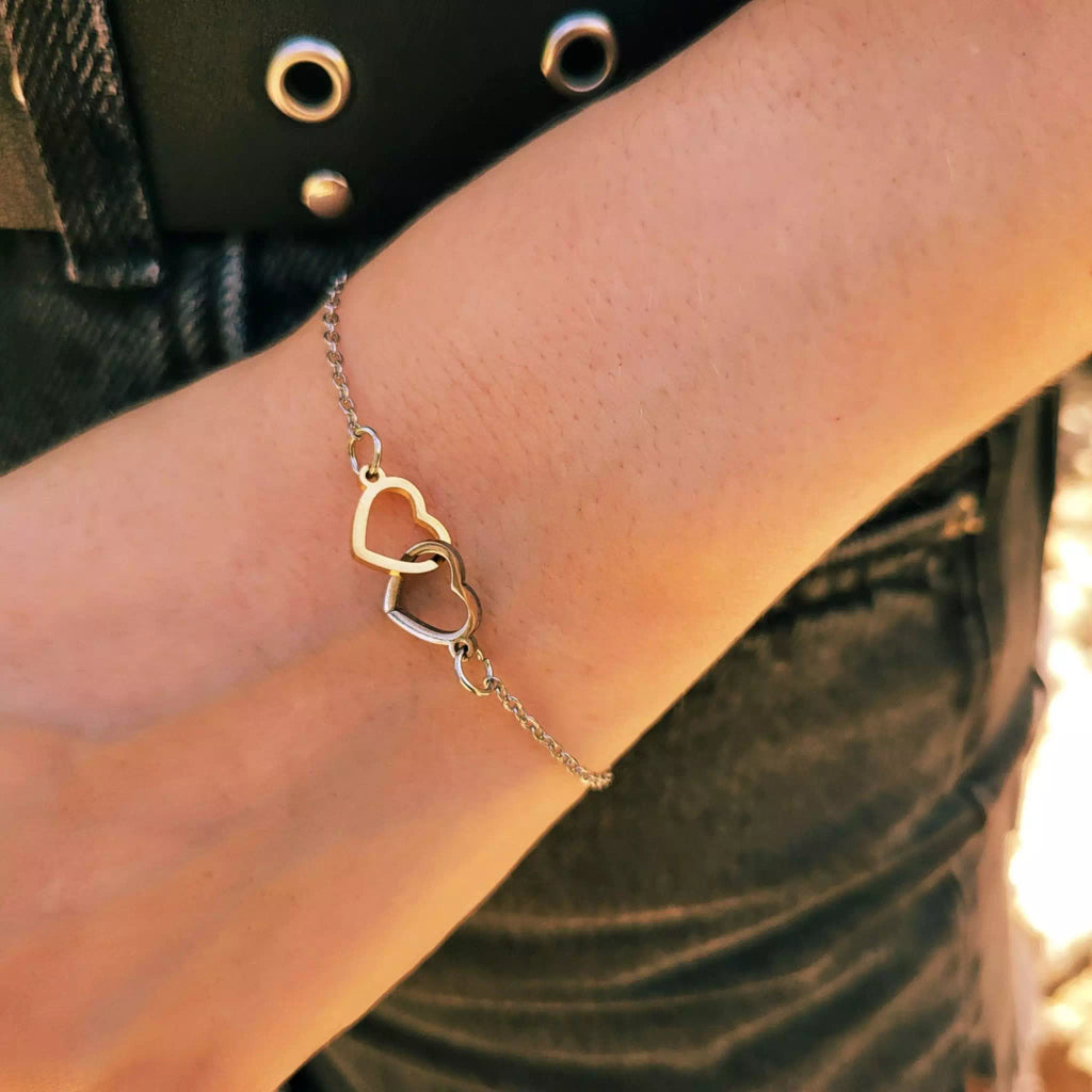 Plan détaillé sous un angle légèrement différent du bracelet bicolore, mettant l'accent sur les formes de coeurs évidées or et argent entrelacées et la fine chaine de liaison. Le bracelet repose sur un poignet, avec le bord d'une ceinture noire à œillets et un jean gris foncé visible en arrière-plan.
