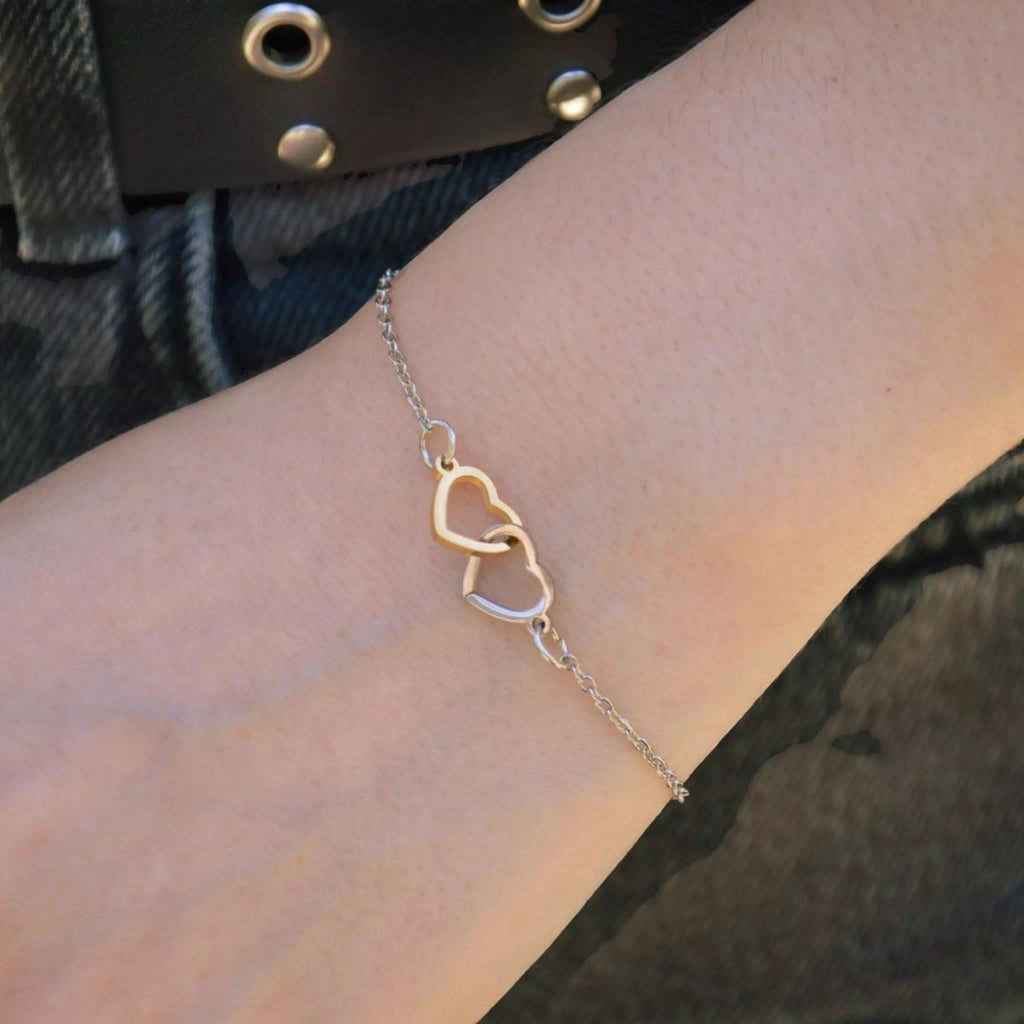 Plan de face du poignet d'une personne portant le bracelet chaine bicolore avec les deux coeurs évidés or et argent entrelacés. Le poignet est positionné devant un jean en denim gris foncé avec le détail d'une ceinture noire à œillets.