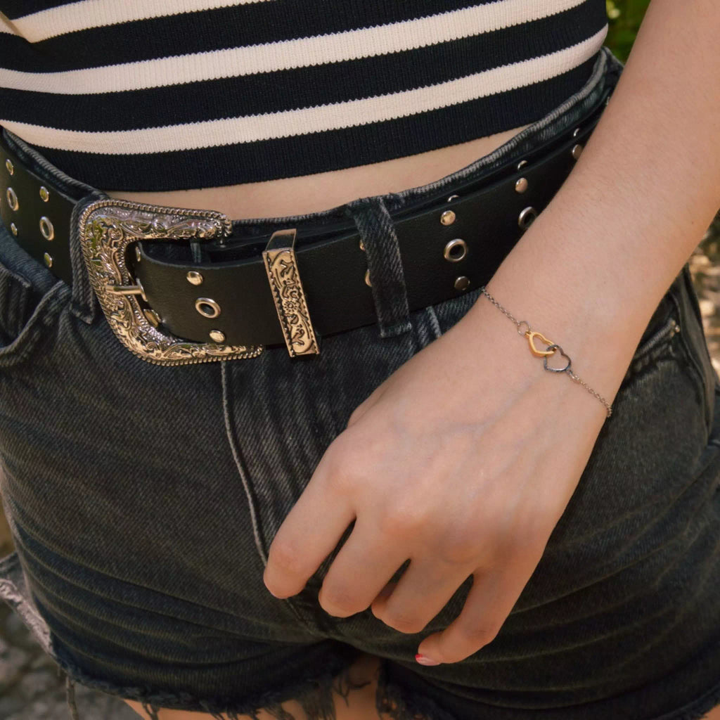 Une vue de profil à mi-corps d'une personne portant le bracelet bicolore à deux coeurs entrelacés à son poignet. La main est détendue sur le côté de la cuisse, vêtue d'un short en jean gris foncé déchiqueté, d'une ceinture en cuir noir ornée d'une boucle gravée et d'un haut à rayures horizontales noires et blanches. L'arrière-plan montre une texture pavée et de la verdure floue.