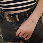 Une vue de profil à mi-corps d'une personne portant le bracelet bicolore à deux coeurs entrelacés à son poignet. La main est détendue sur le côté de la cuisse, vêtue d'un short en jean gris foncé déchiqueté, d'une ceinture en cuir noir ornée d'une boucle gravée et d'un haut à rayures horizontales noires et blanches. L'arrière-plan montre une texture pavée et de la verdure floue.