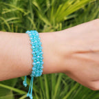 Vue latérale d'un bracelet artisanal bleu ciel en perles de cristal et macramé, porté sur un bras. Le tissage complexe et géométrique met en valeur la brillance des perles facettées sous une lumière naturelle. L'arrière-plan montre un feuillage tropical d'un vert vif.