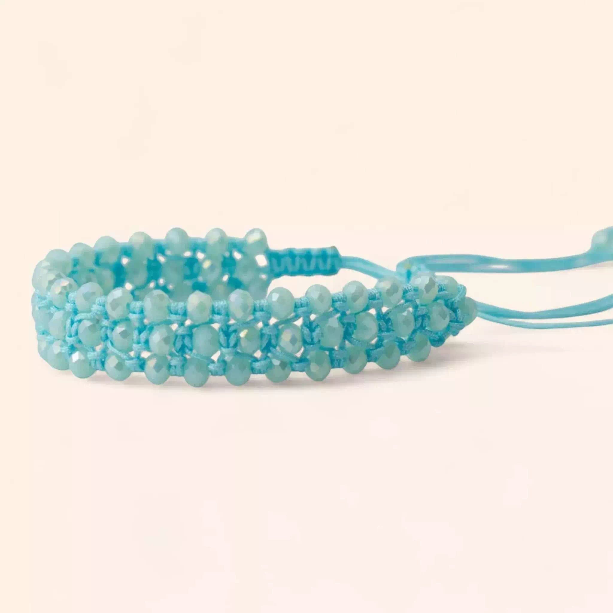 Vue de profil du bracelet en perles bleu ciel et fils de macramé tressés, disposé horizontalement sur un fond neutre beige clair. On aperçoit distinctement la finition de la fermeture et les cordons de serrage sur le côté droit.