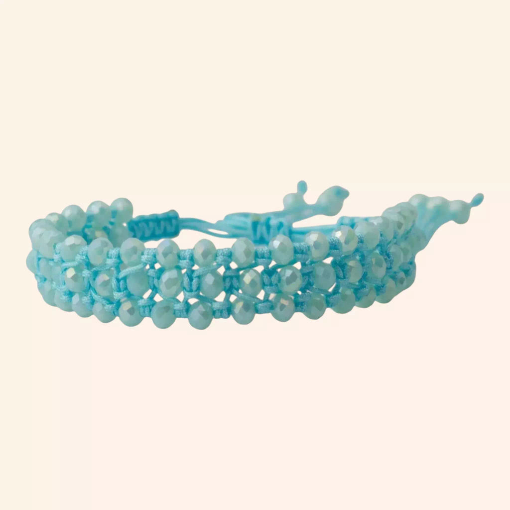Présentation du bracelet bleu ciel en cristal et macramé sous un angle de trois-quarts, isolé sur un fond uni beige. Cette vue met en évidence l'épaisseur, la texture du tissage et la fermeture réglable située à l'arrière.