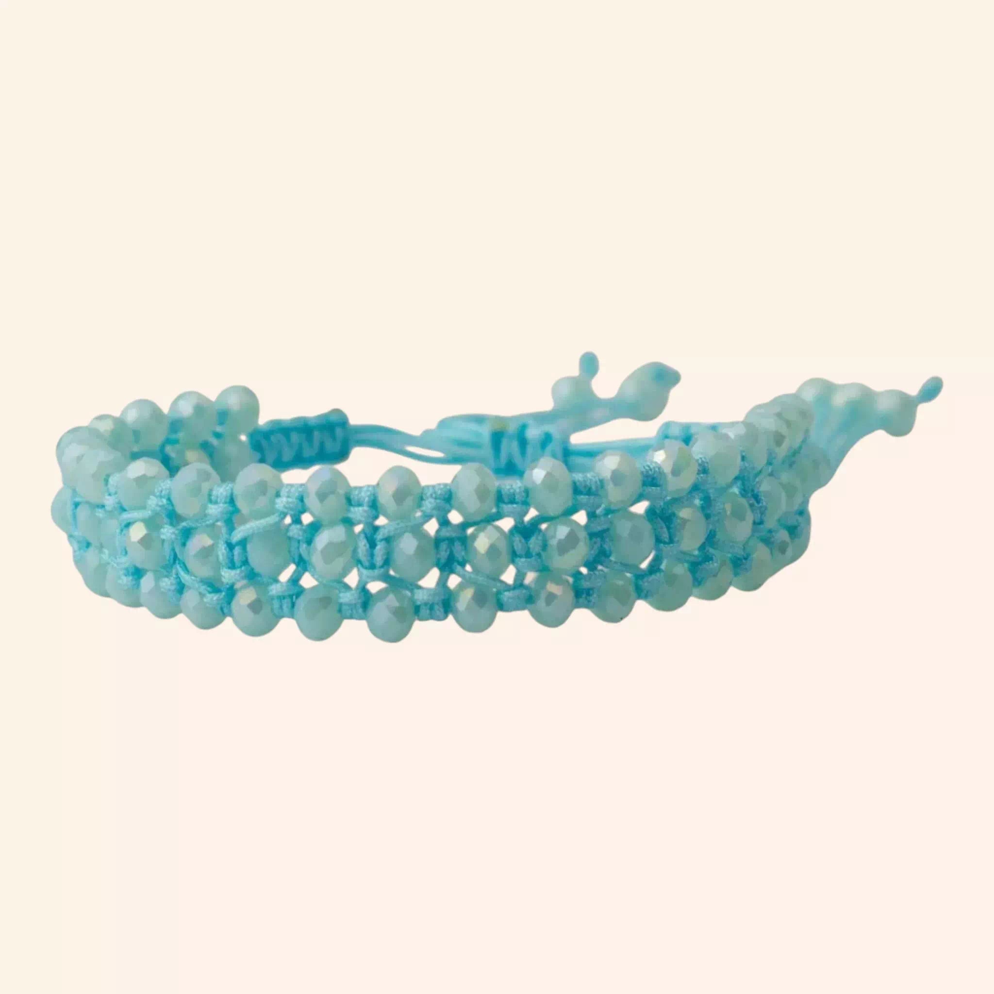 Présentation du bracelet bleu ciel en cristal et macramé sous un angle de trois-quarts, isolé sur un fond uni beige. Cette vue met en évidence l'épaisseur, la texture du tissage et la fermeture réglable située à l'arrière.