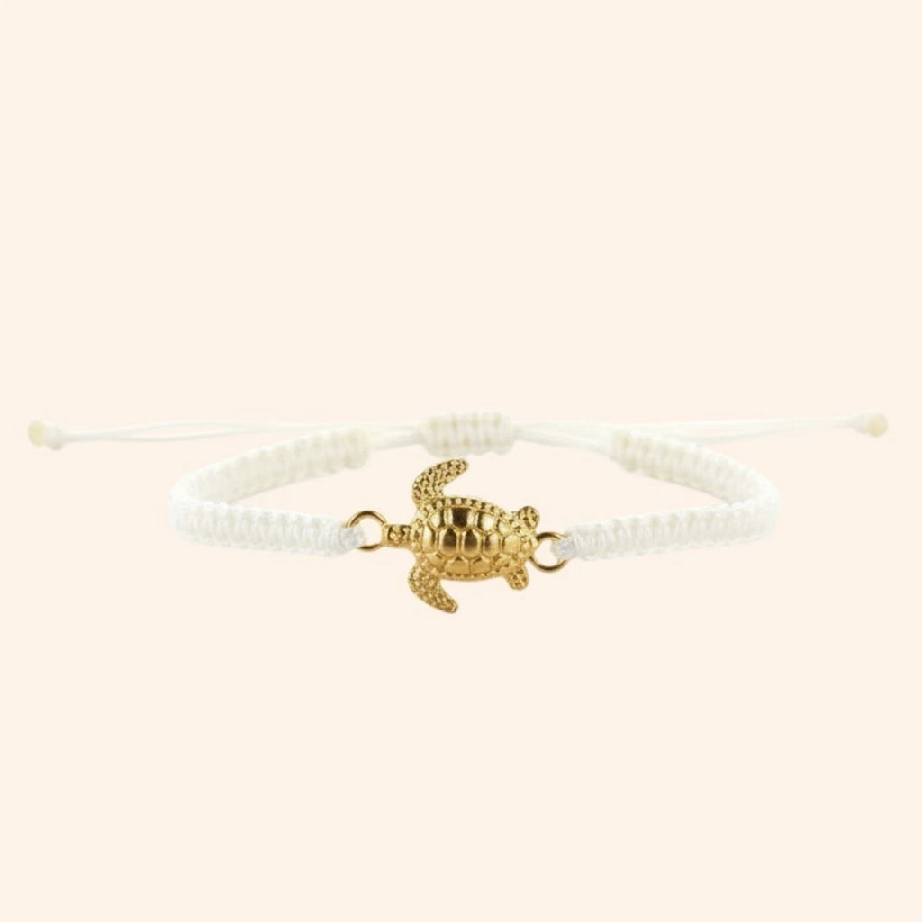 Vue de face d'un bracelet en macramé blanc avec un pendentif central en forme de tortue dorée sur fond beige clair.