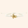 Vue de face d'un bracelet en macramé blanc avec un pendentif central en forme de tortue dorée sur fond beige clair.