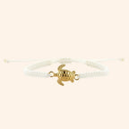 Vue de face d'un bracelet en macramé blanc avec un pendentif central en forme de tortue dorée sur fond beige clair.