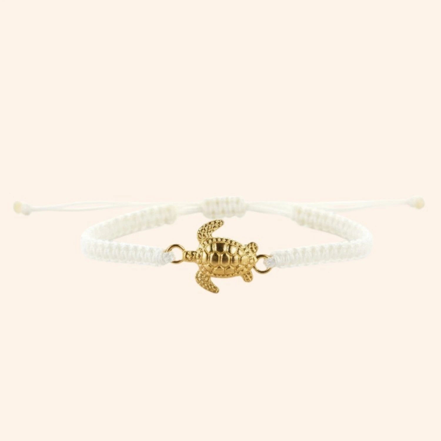 Vue de face d'un bracelet en macramé blanc avec un pendentif central en forme de tortue dorée sur fond beige clair.