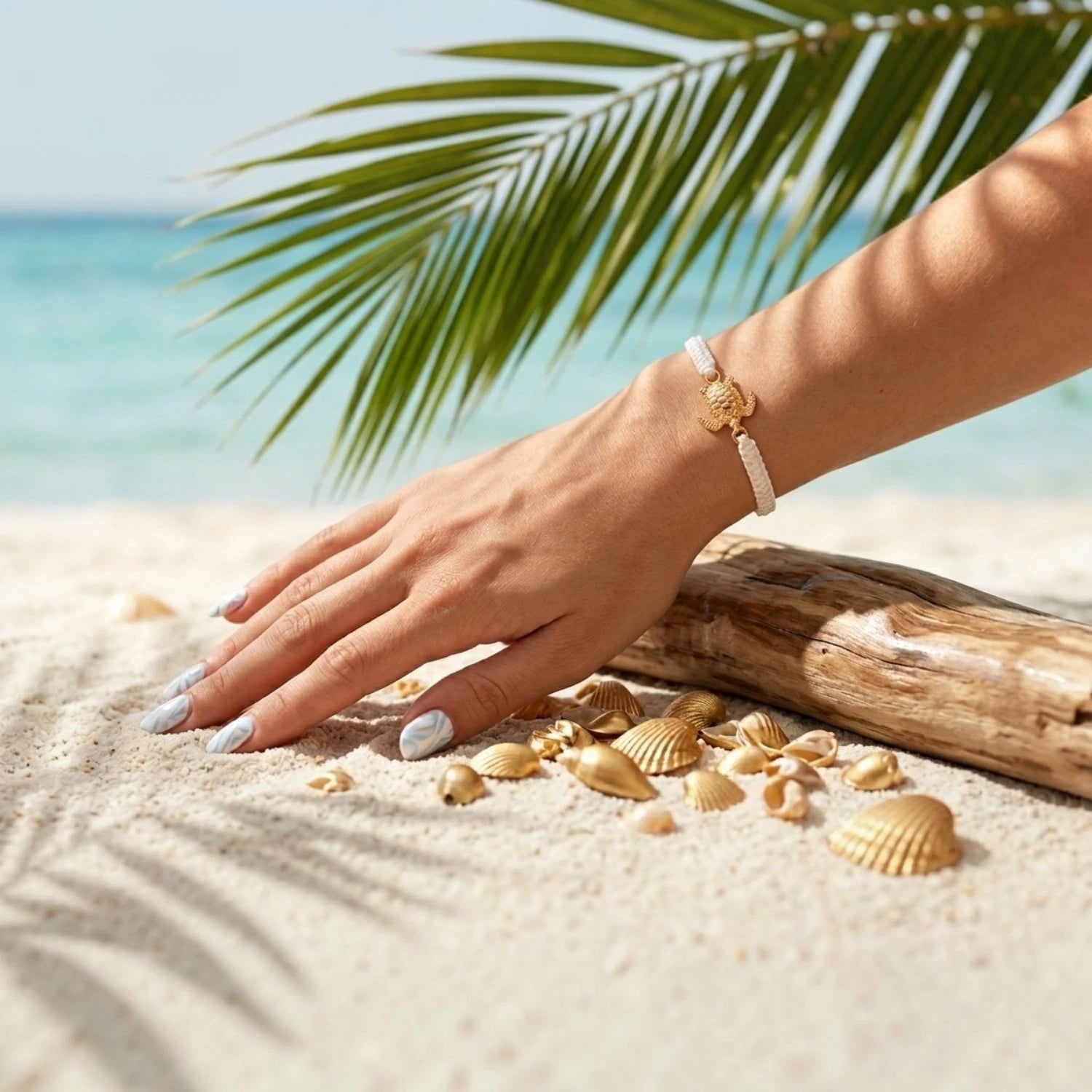 Mise en scène estivale d'un bracelet en macramé blanc orné d'une tortue dorée, porté au poignet sur une plage de sable fin avec la mer turquoise et une feuille de palmier en arrière-plan.