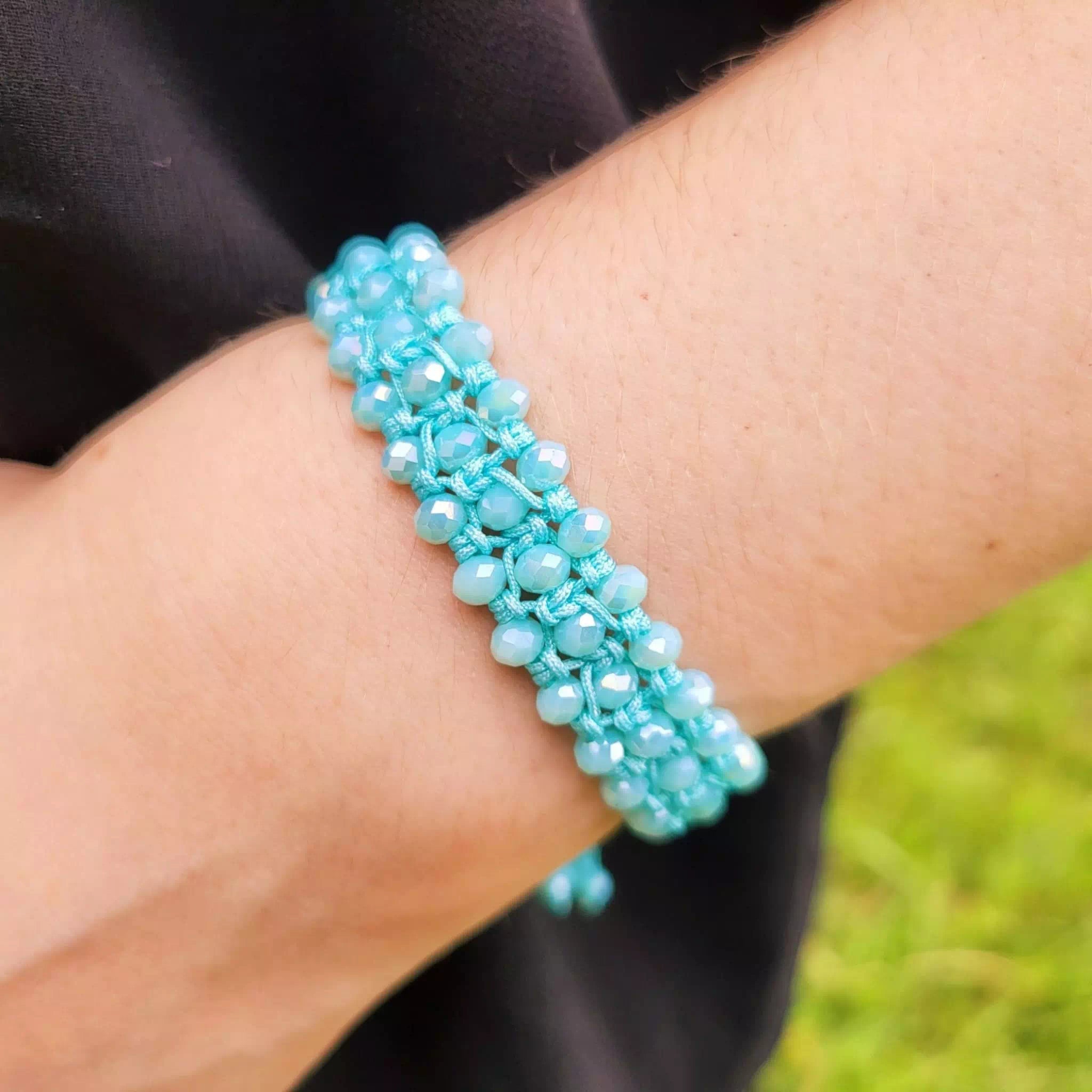 Gros plan du poignet d'une personne portant un bracelet en macramé bleu ciel. Le bijou est composé de trois rangées de perles de cristal bleu clair facettées, tissées ensemble avec un fil assorti. La personne porte un vêtement noir, et l'arrière-plan montre de l'herbe verte floue.