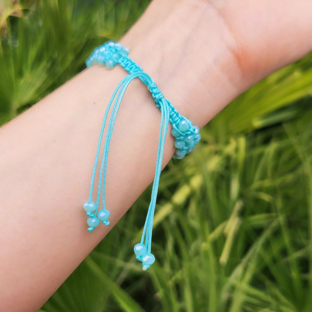 Zoom sur le système de fermeture réglable d'un bracelet en macramé bleu ciel porté au poignet. On distingue le nœud coulissant central et les fils qui pendent, chacun se terminant par de petites perles bleues suspendues. L'arrière-plan est un fond de feuilles vertes floues.