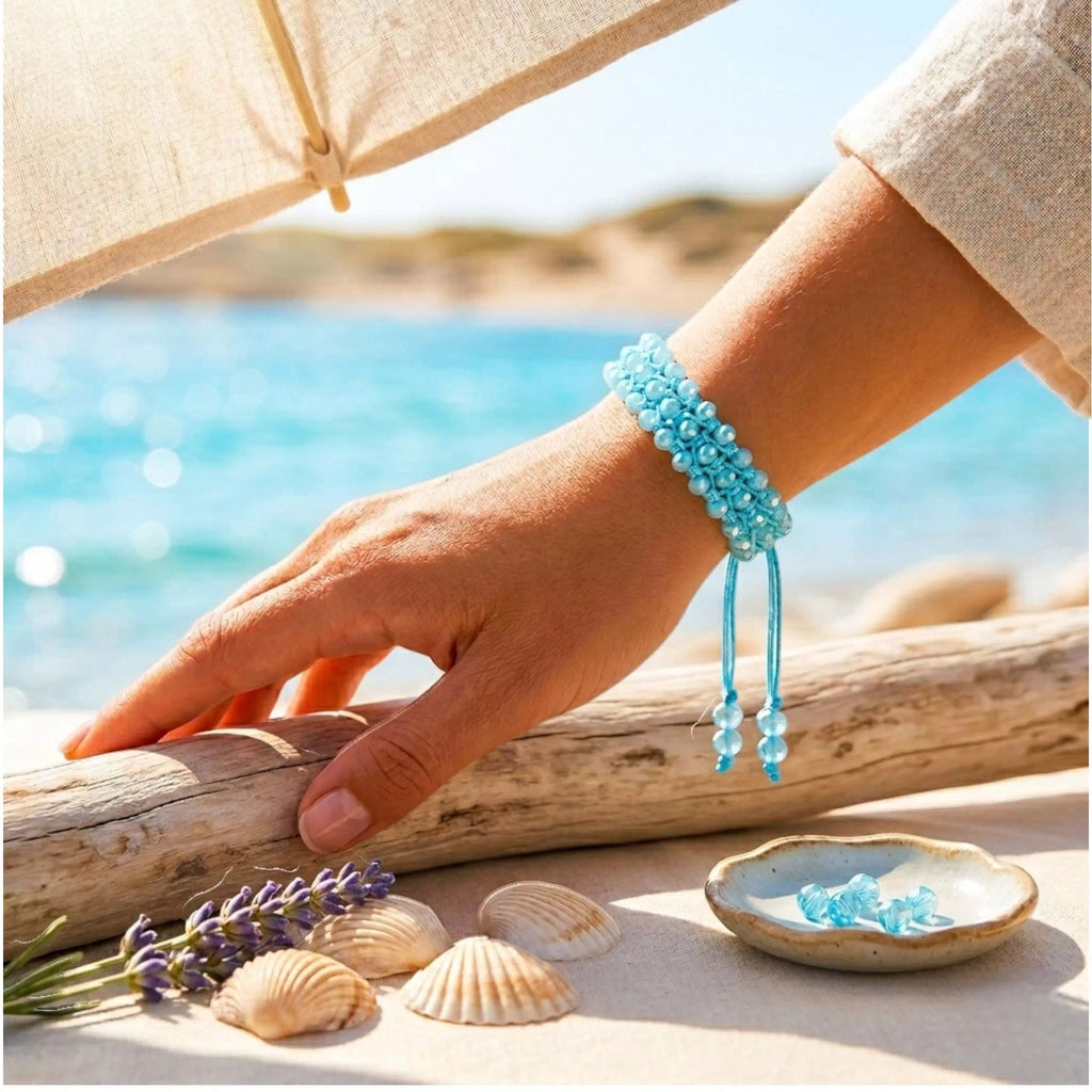 Mise en scène estivale et lumineuse d'un bras posé sur un morceau de bois flotté au bord de la mer, arborant le bracelet en macramé bleu ciel. Au premier plan, on trouve des coquillages, un brin de lavande et une petite coupelle contenant des perles bleues. L'arrière-plan dévoile une plage ensoleillée et une mer turquoise scintillante sous un parasol en toile.