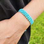 Vue rapprochée d'un bracelet manchette en macramé bleu ciel porté au poignet. Le bracelet, large et structuré, est orné de multiples perles en cristal bleu étincelantes intégrées dans un tissage de nœuds fins. L'arrière-plan se compose d'un tissu noir et d'une pelouse verte en flou artistique.