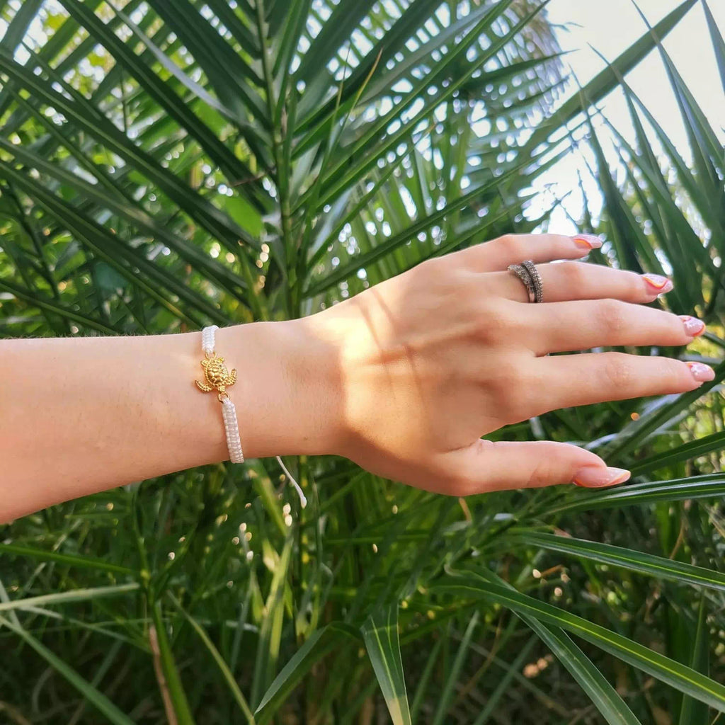 Bracelet en cordon tressé blanc orné d'une tortue dorée, porté au poignet d'une femme devant des feuilles de palmier.