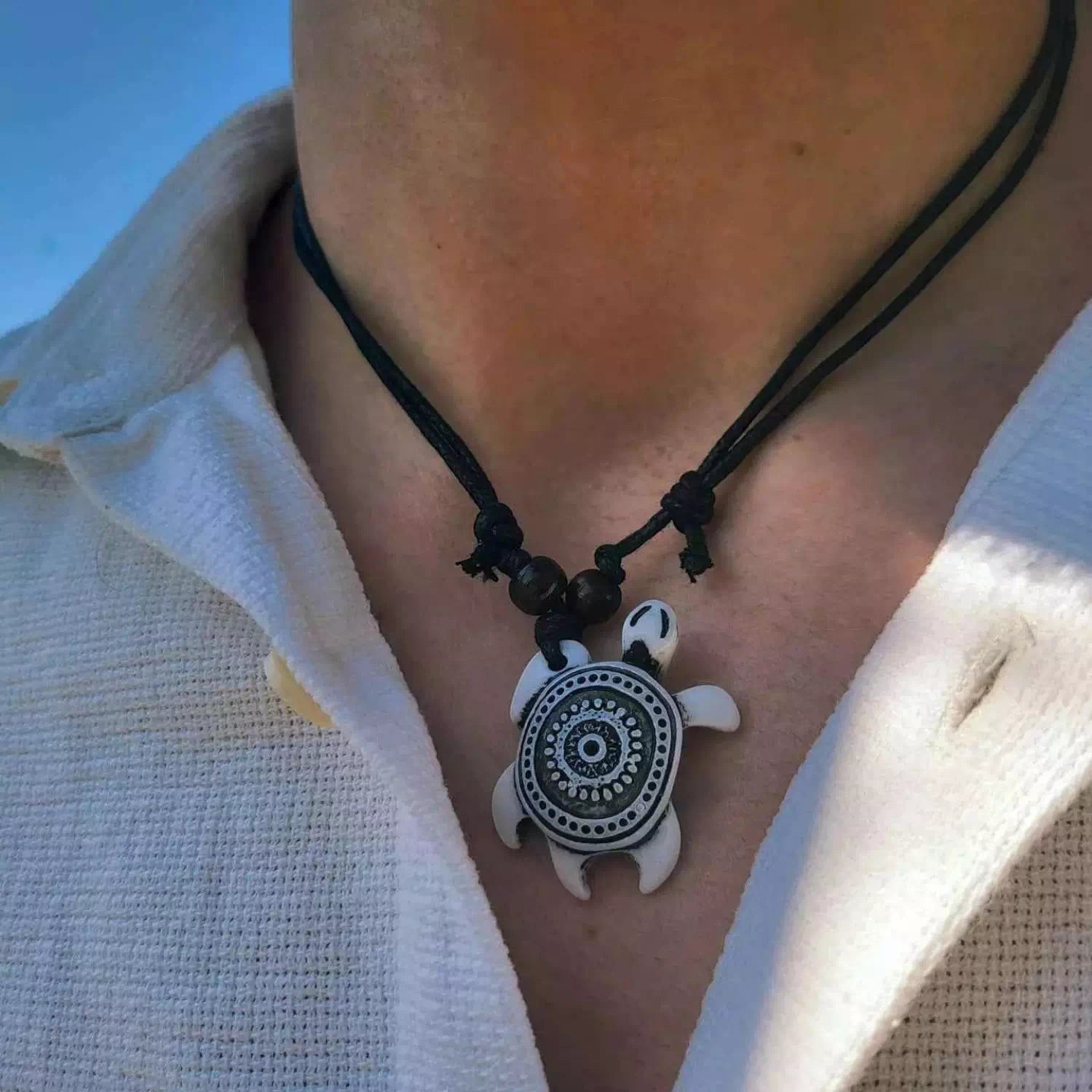 Zoom sur un homme portant le collier de style surfeur avec son pendentif tortue blanche et noire sur une chemise blanche.