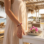 Photo style de vie d'une femme en robe d'été en lin beige portant le bracelet œil de tigre rose Jethyka, accoudée à une table près de la mer.